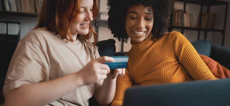 Two young women shopping online from the laptop sitting on the couch. Interracial couple of girlfriends using credit card to shop from an e-commerce website.