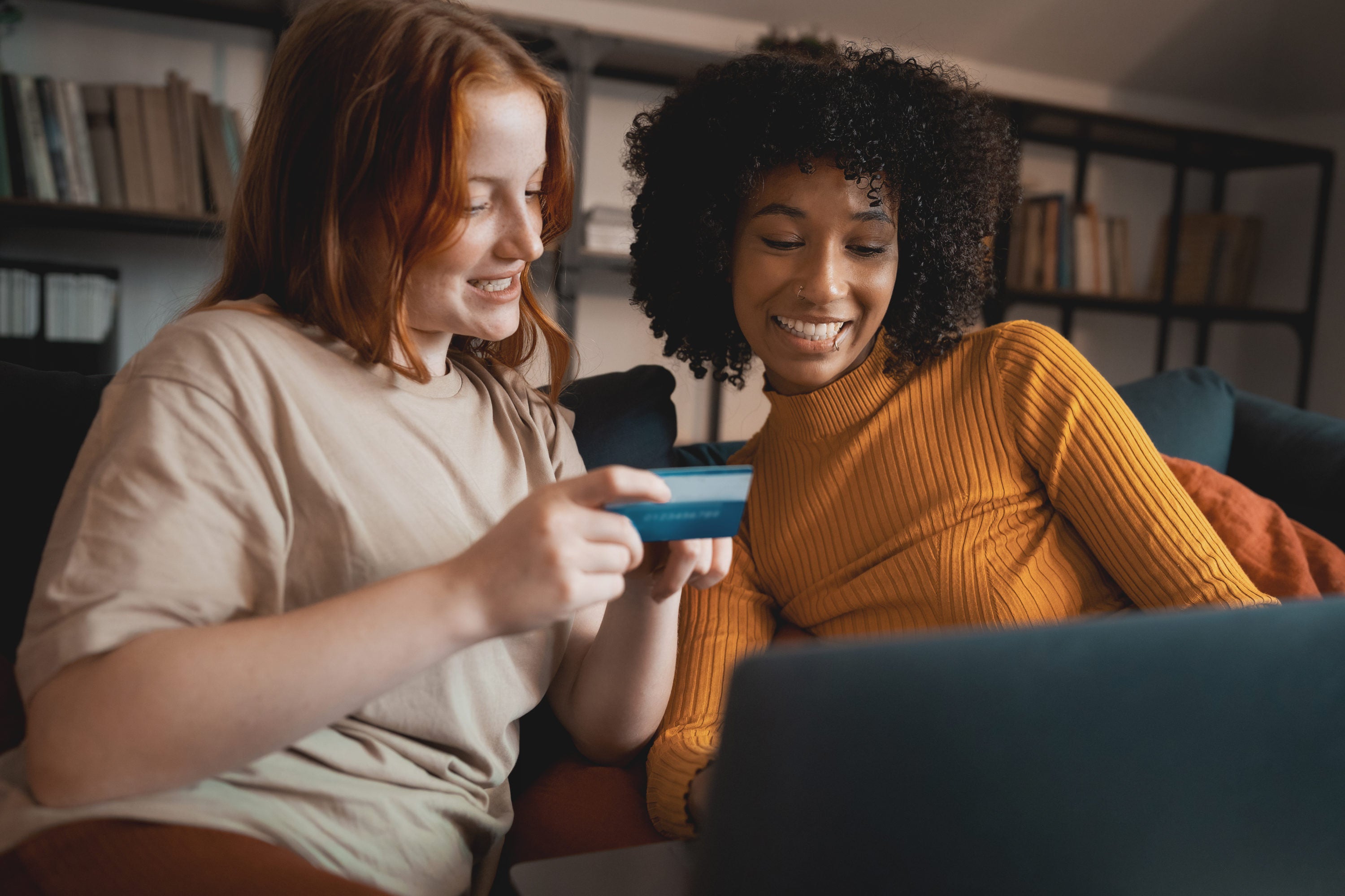 Two young women shopping online from the laptop sitting on the couch. Interracial couple of girlfriends using credit card to shop from an e-commerce website.