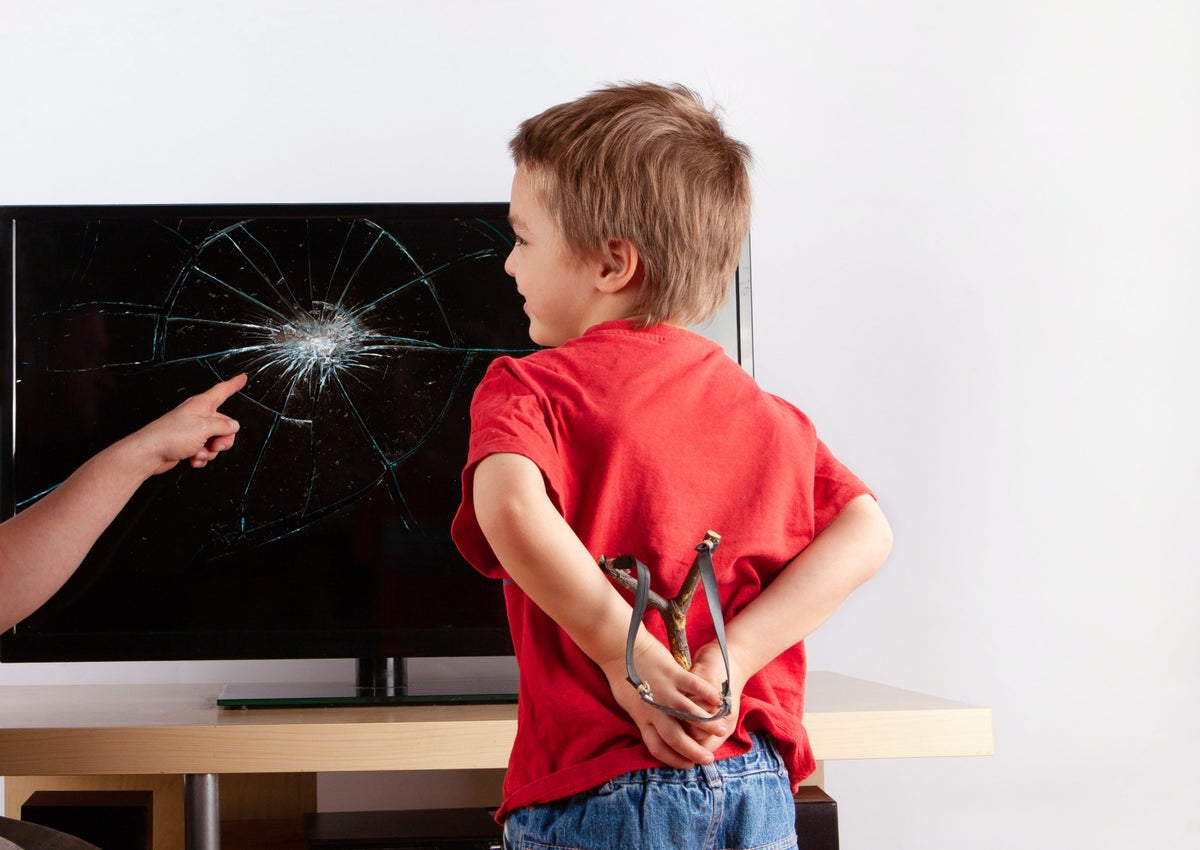 Little boy standing in front of a TV with broken screen and hiding a slingshot behind his back while a hand pointing to the broken display