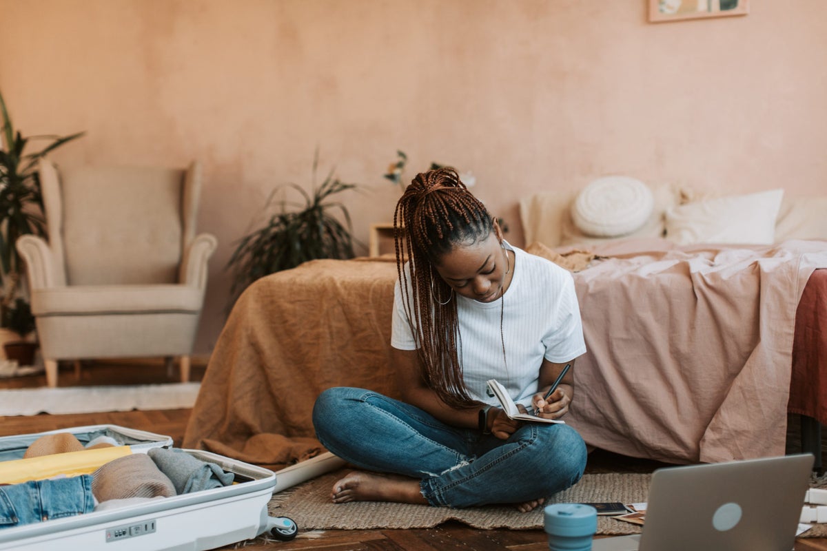 Woman writing a packing list while sitting on her bedroom floor in front of her bed. Suitcase is in front of her.