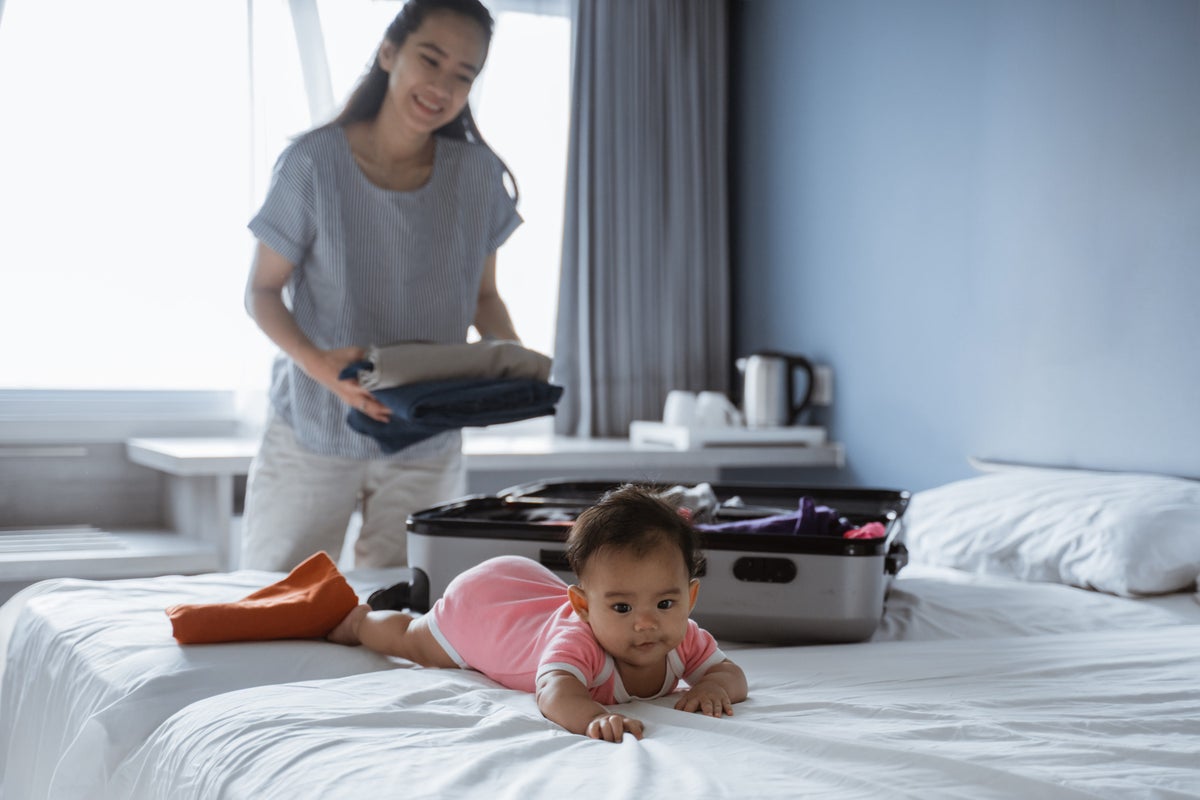 A mother packs a suitcase on the bed. Her baby is on their tummy, also on the bed, smiling at the camera.