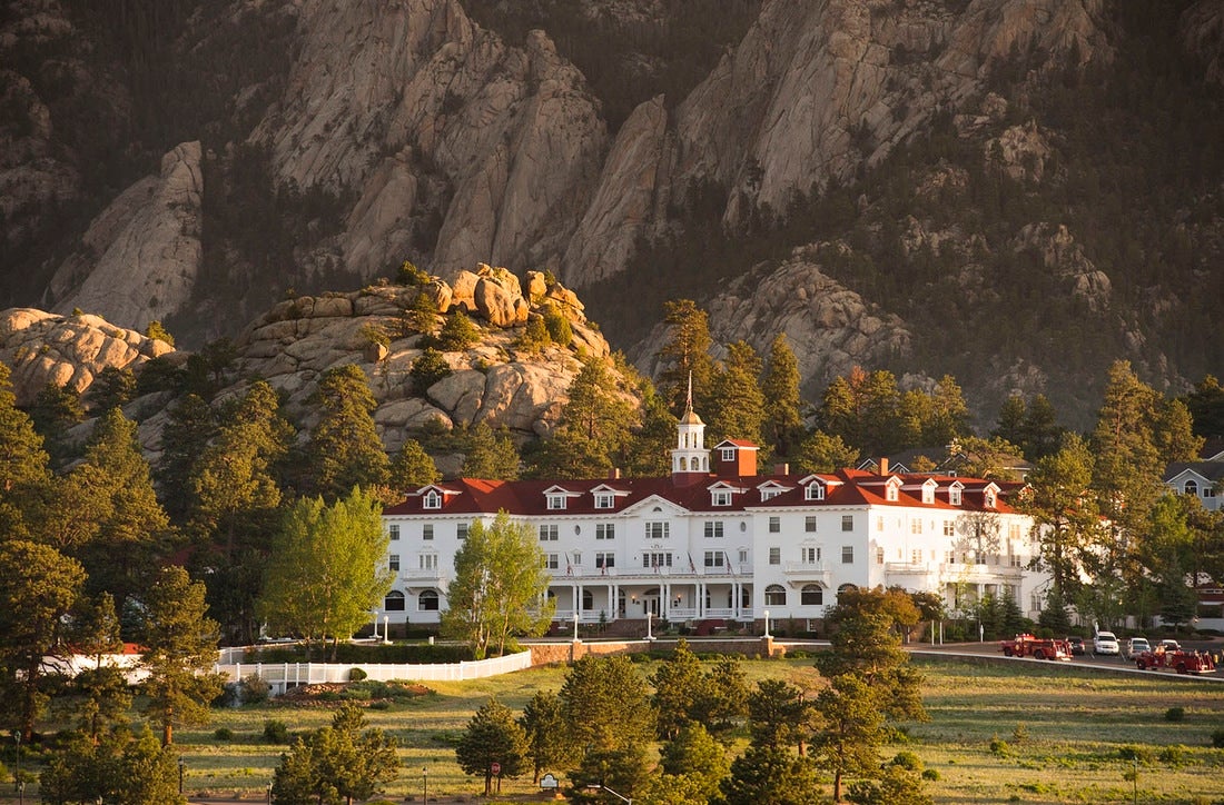 An aerial view The Stanley Hotel in Colorado. The hotel is set in front of mountains with trees in front.