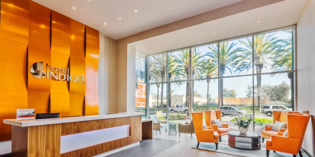 The lobby at Hotel Indigo Anaheim has a bright, orange textured wall behind the hotel lobby with seating to the left in front floor to ceiling windows.