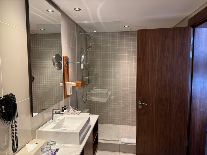 An image from inside of the bathroom with the shower in the background and singel sink in the foreground. The sink has 2 glasses, a tissue box, and full size body lotion and hand soap.